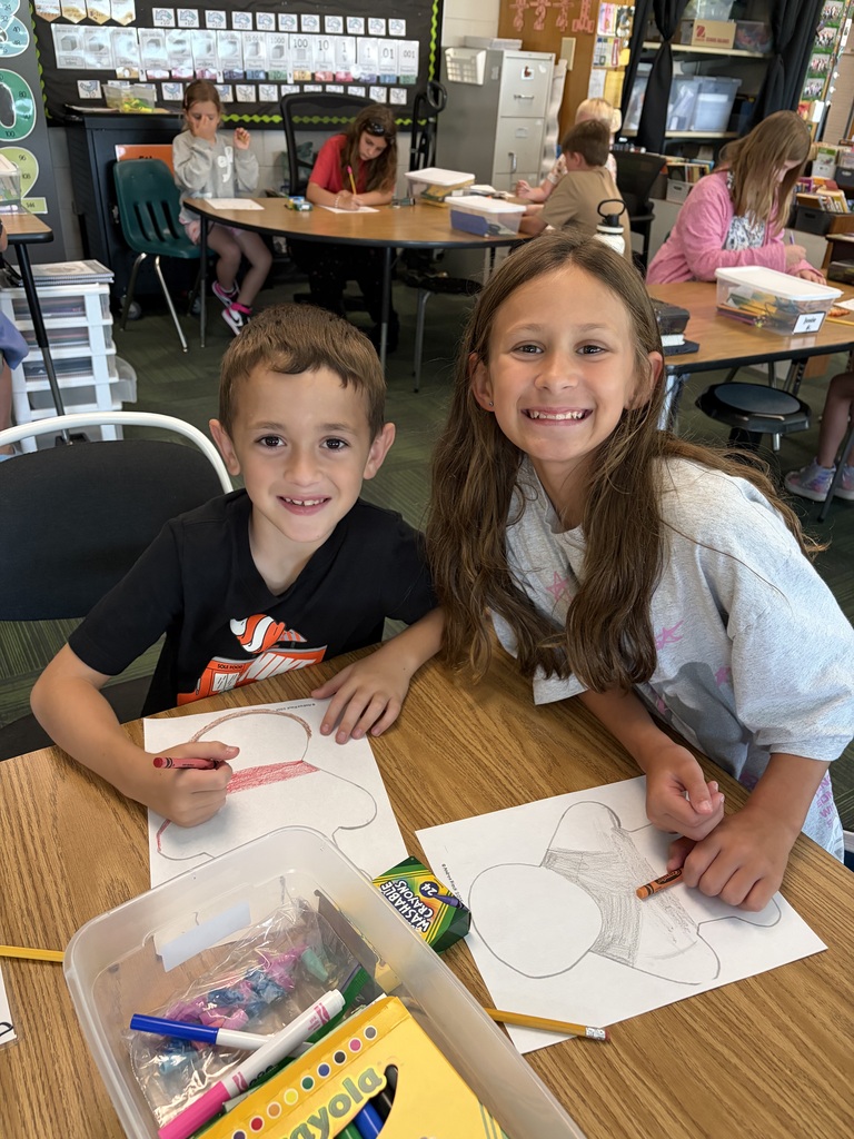 A boy and girl pairing smiling together as they work on outlining their paper figures with crayons at a shared desk.