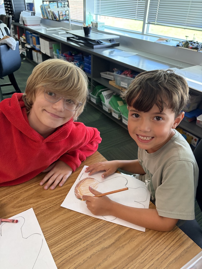 Two boys sitting at a desk together smiling while one uses a colored pencil to detail his project.