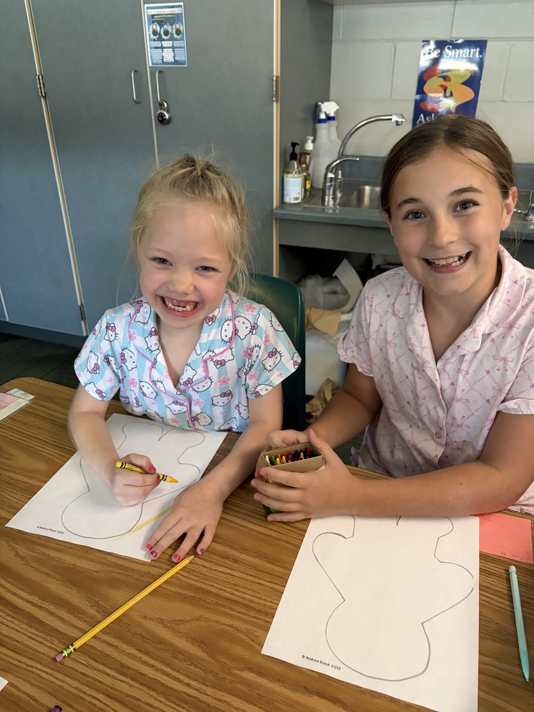 Two smiling girls sitting together at a wooden desk with a crayon box and paper cutouts.