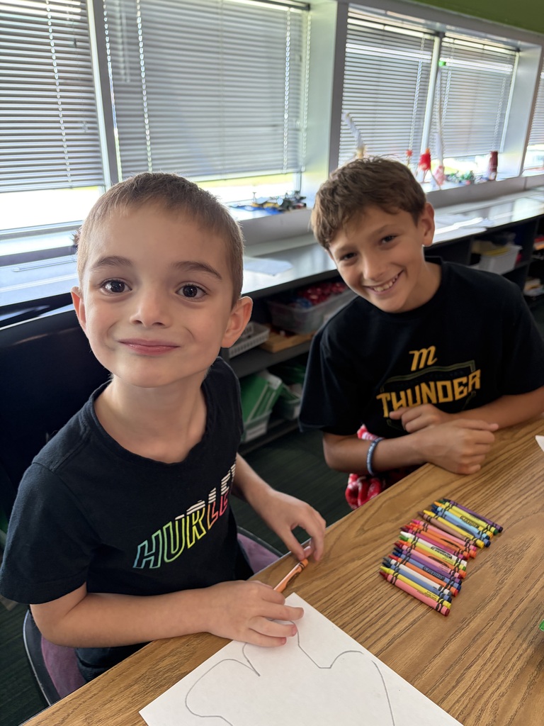 Two boys smiling side-by-side at a wooden table while starting to work on their paper outline coloring project.