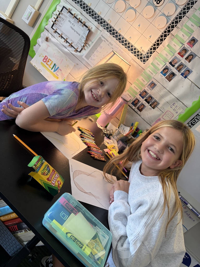 Two girls smiling bright at a table full of crayons while coloring their paper outline projects.