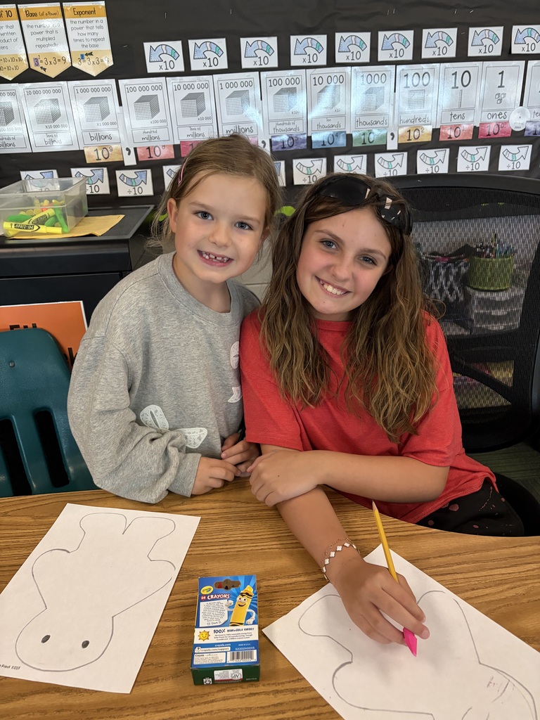 Two girls leaning together and smiling while using crayons on their paper figure outlines.