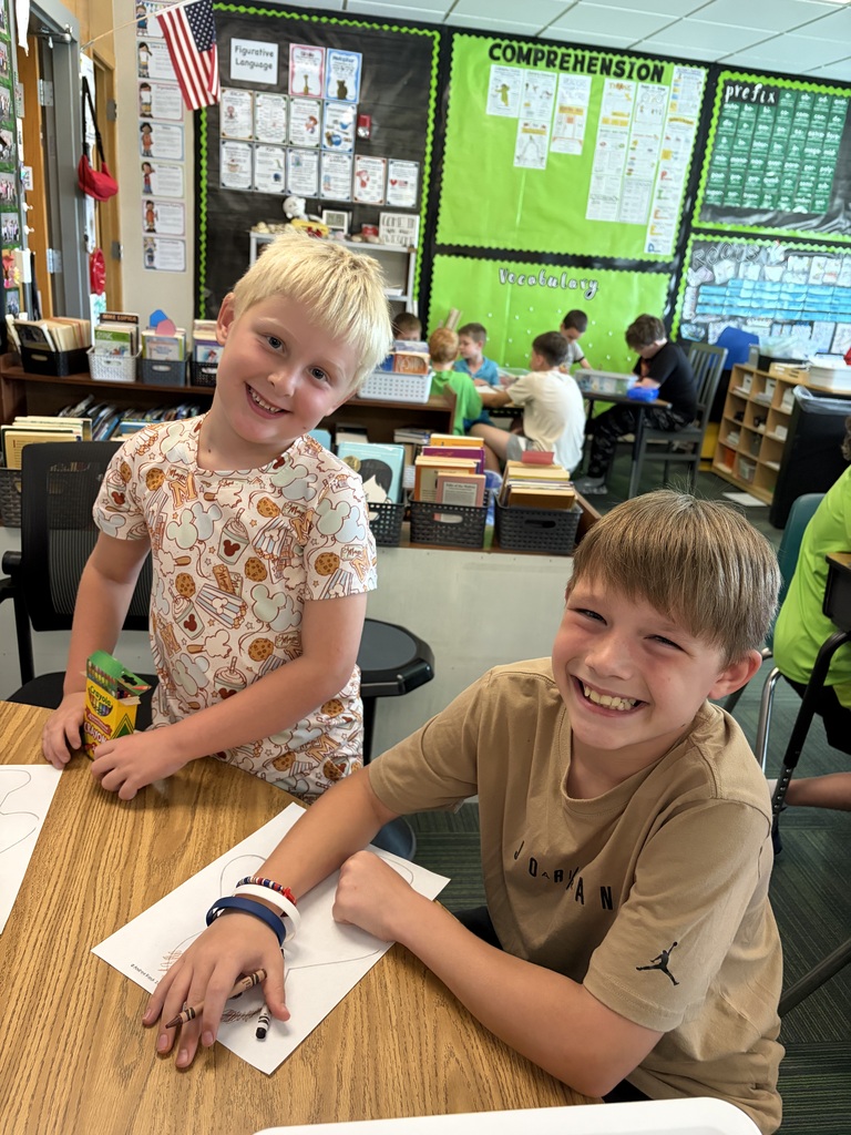 Two smiling boys sitting at a desk with crayons and their paper figure coloring sheets.