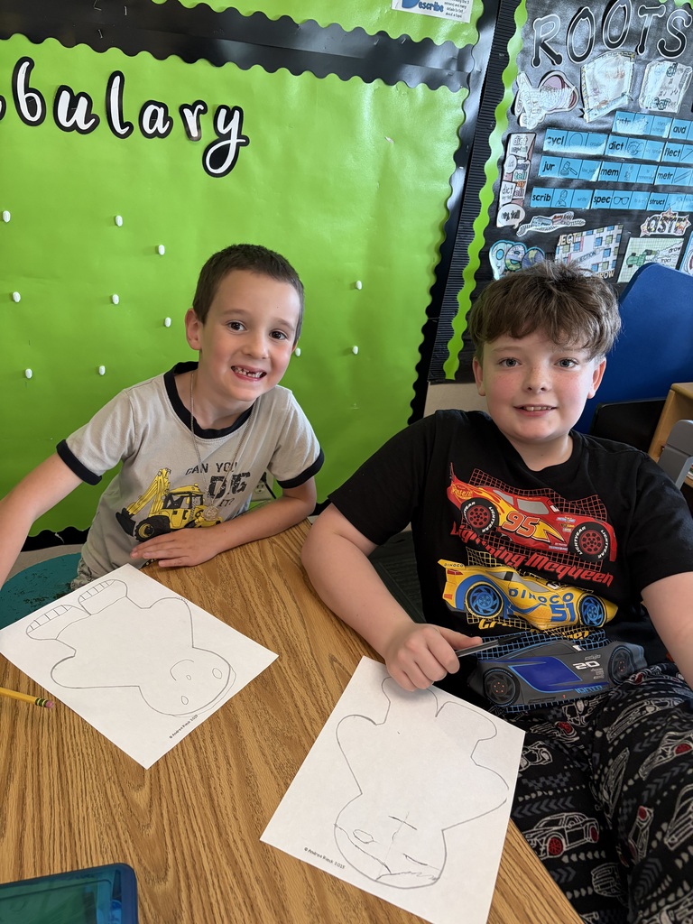 Two boys smiling at the camera while working on their paper figure drawing worksheets at a classroom desk.