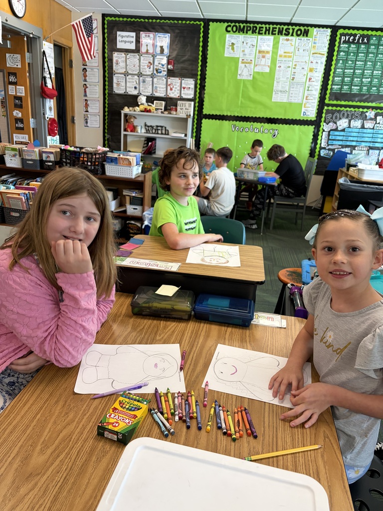 Two girls sitting at a classroom table with crayons, smiling as they work on their paper figure drawings.