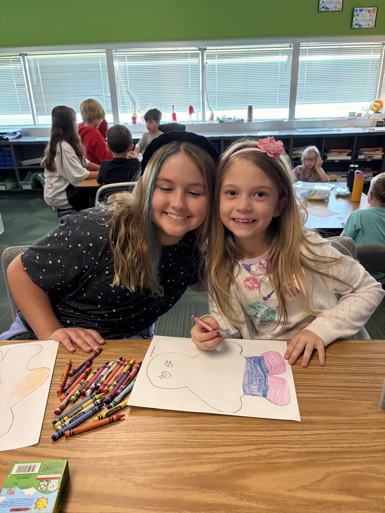 Two girls posing together with warm smiles while working with crayons on a paper figure drawing.