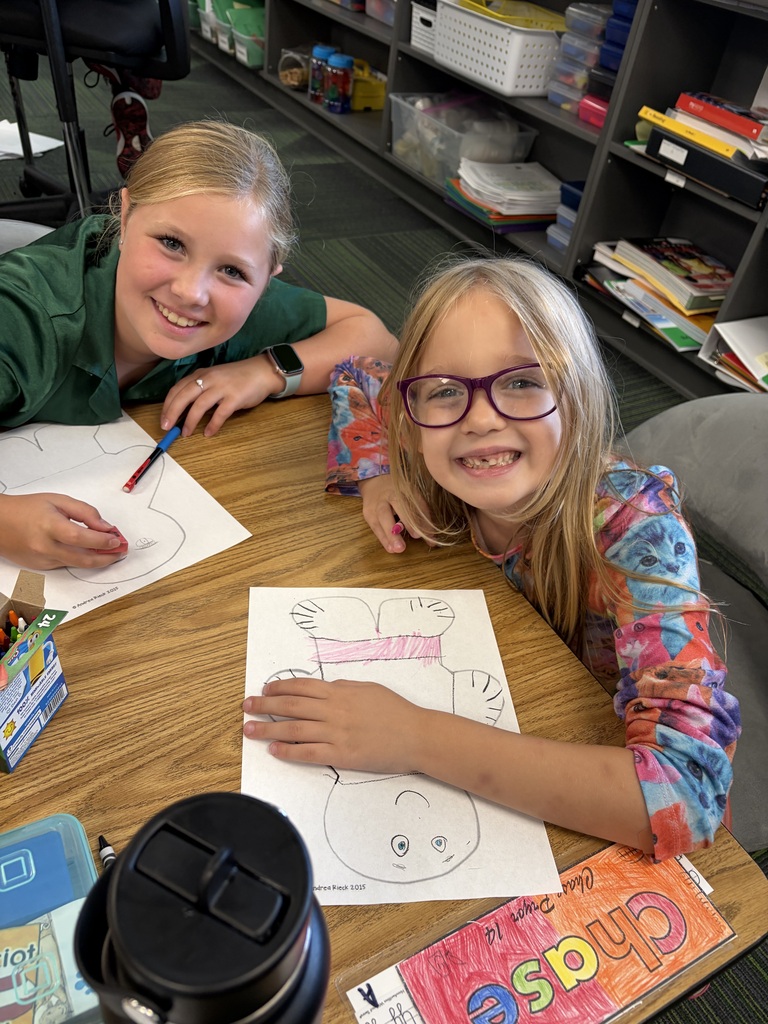 Two girls smiling together at a desk while filling in details on their paper figure sheets.