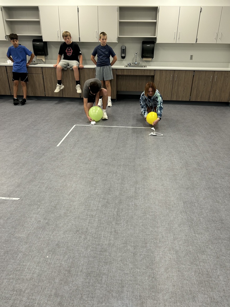 A row of students watch two students on the ground get their wind puft cars ready to race. 