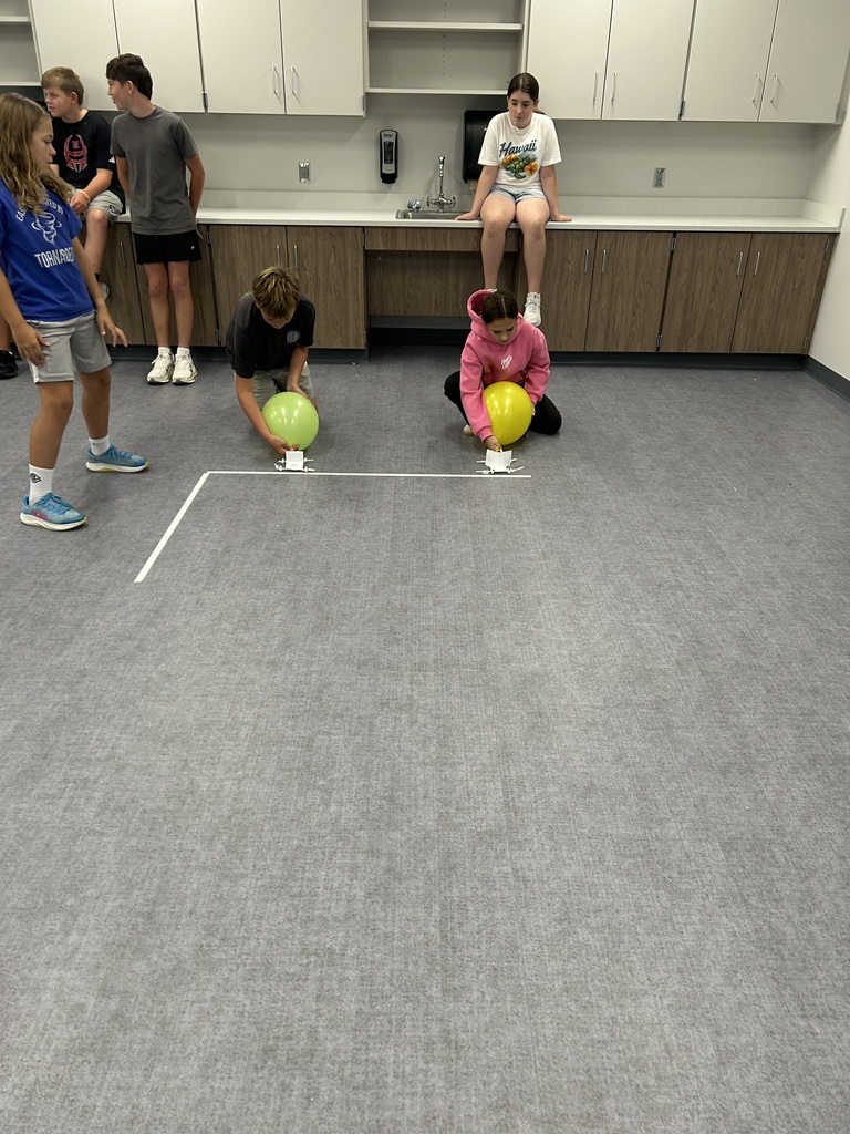 A row of students watch two other students on the ground get their wind puft cars ready to race. 