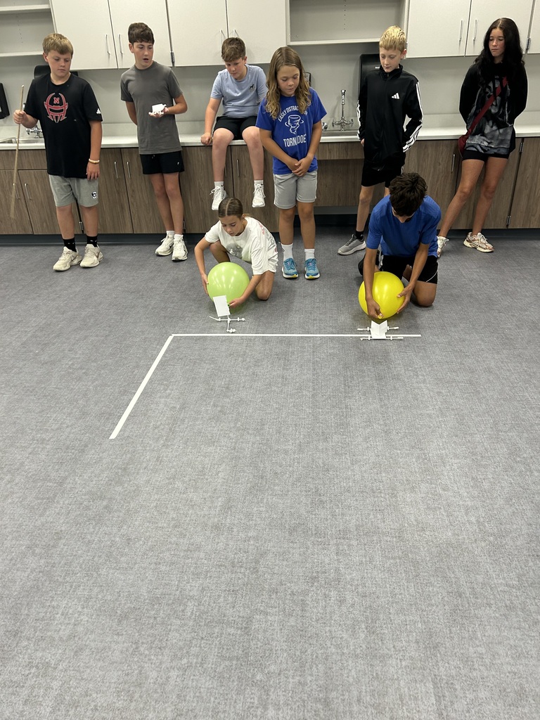 A row of students are watching two other students on the ground get their wind puft cars ready to race. 