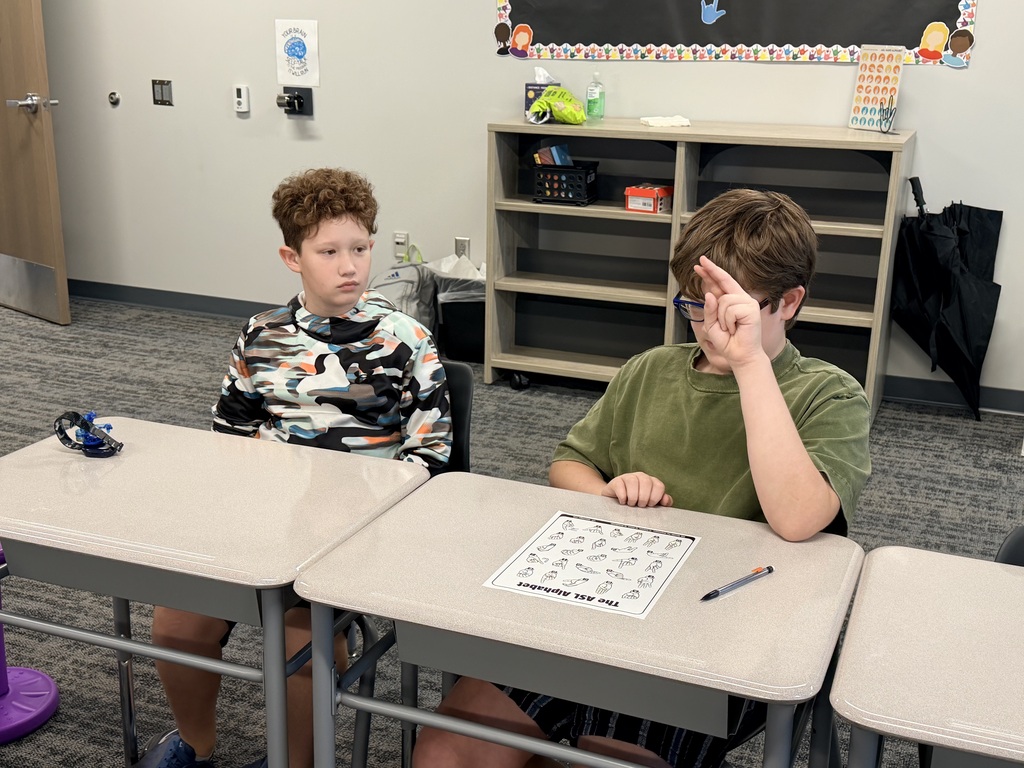 Two students sit at desks and practice ASL. 