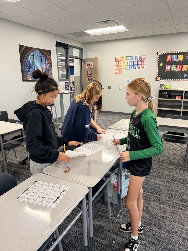 Students are standing around desks looking at pieces of paper. 