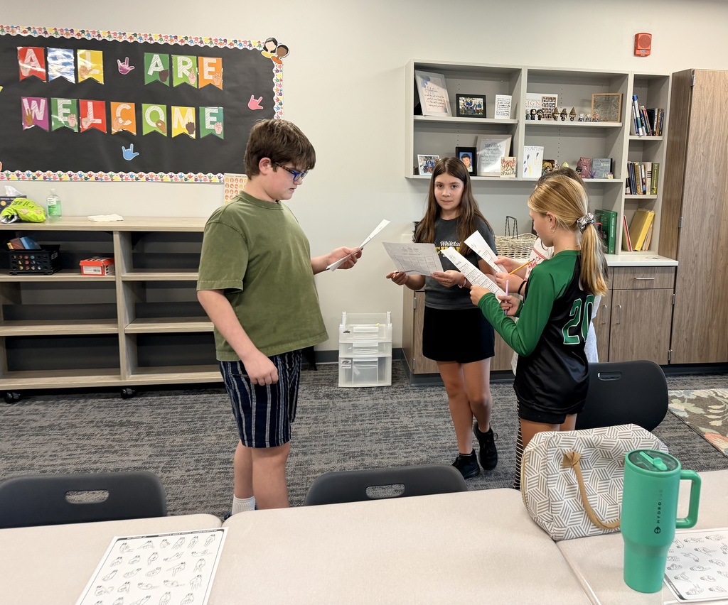 A group of three students is standing together holding worksheets and practicing ASL. 