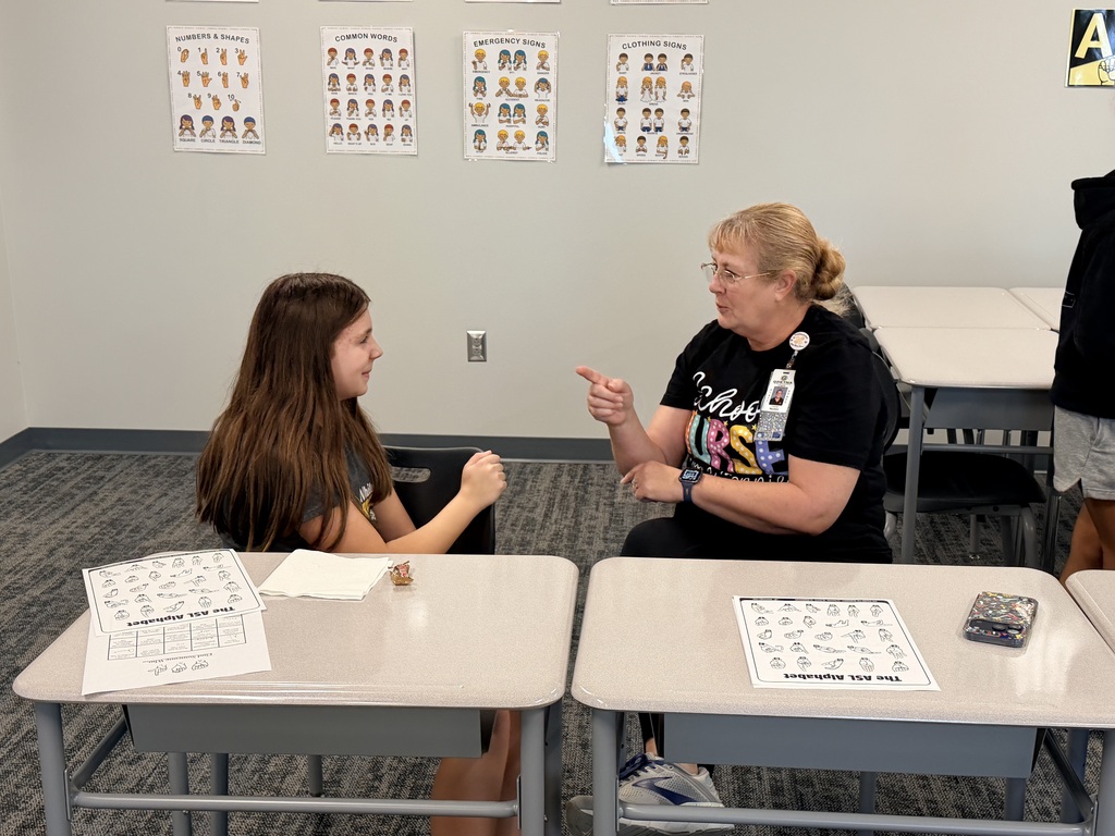 A student and a teacher sit at desks and practice ASL. 