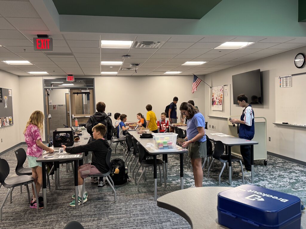 A large group of students stand in a classroom space. They are working on puzzles, crafts, and chess games. 