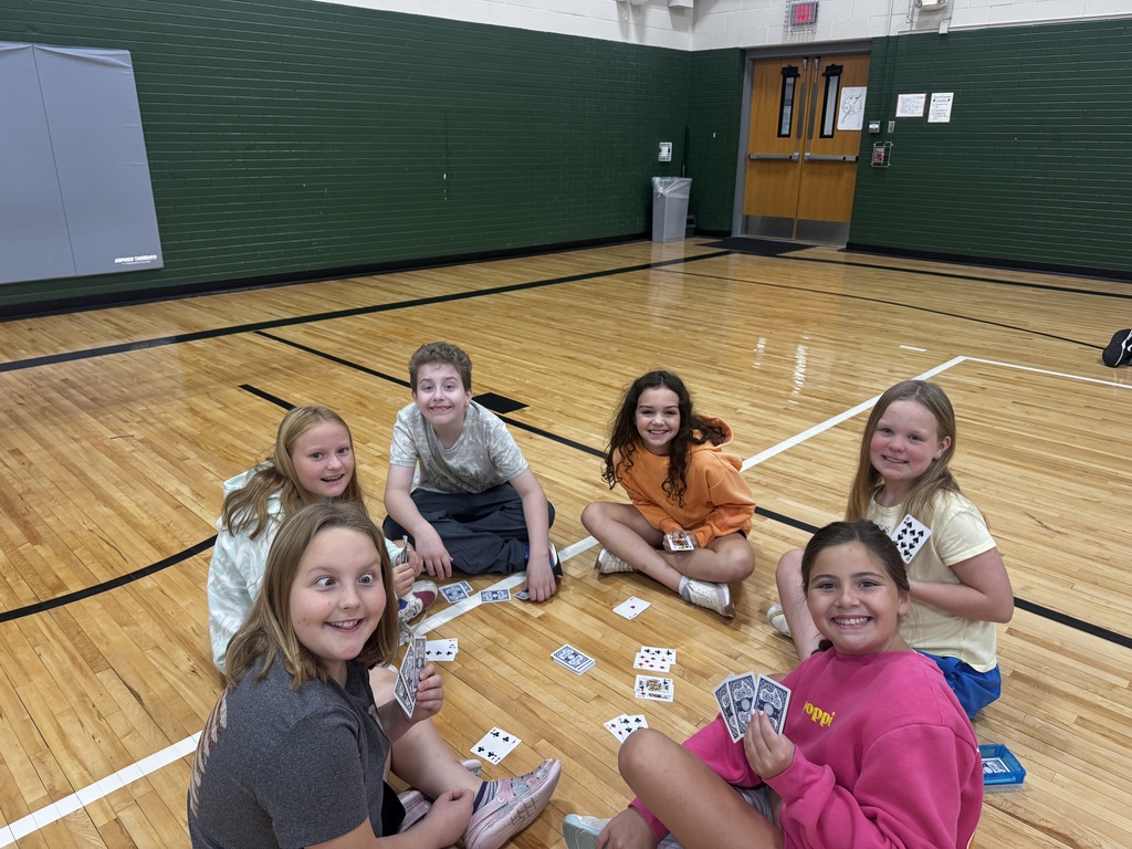 A group of six students sitting on the gym floor smiling around their card game layout.