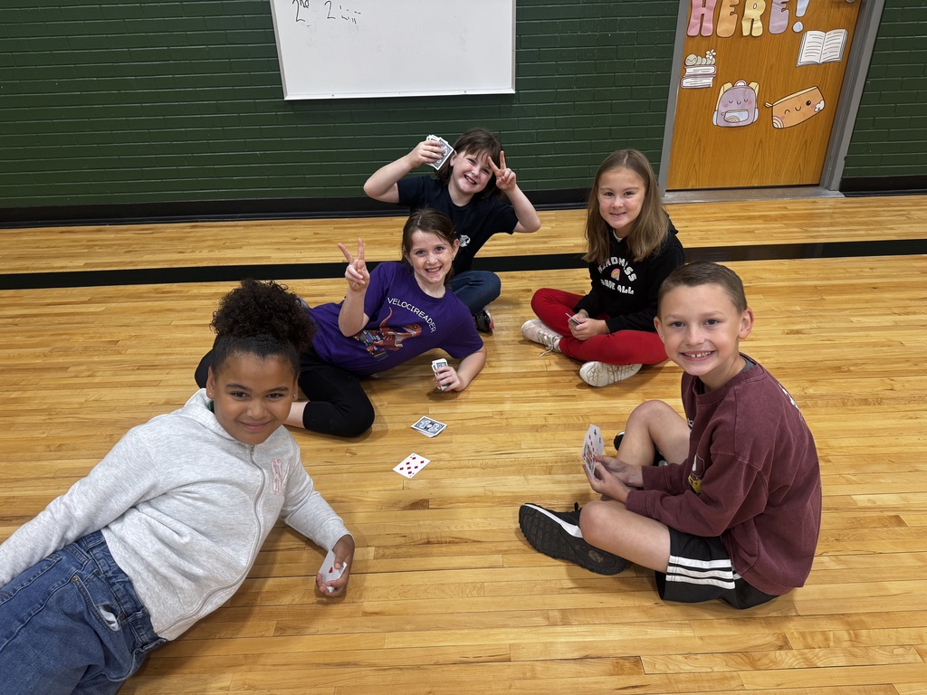 Five students sitting in a circle on the wooden floor holding and playing cards.