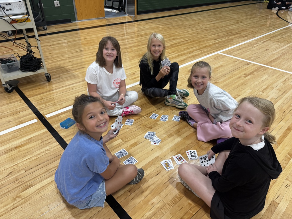 Five young girls sitting on the gymnasium floor smiling while playing a card game.