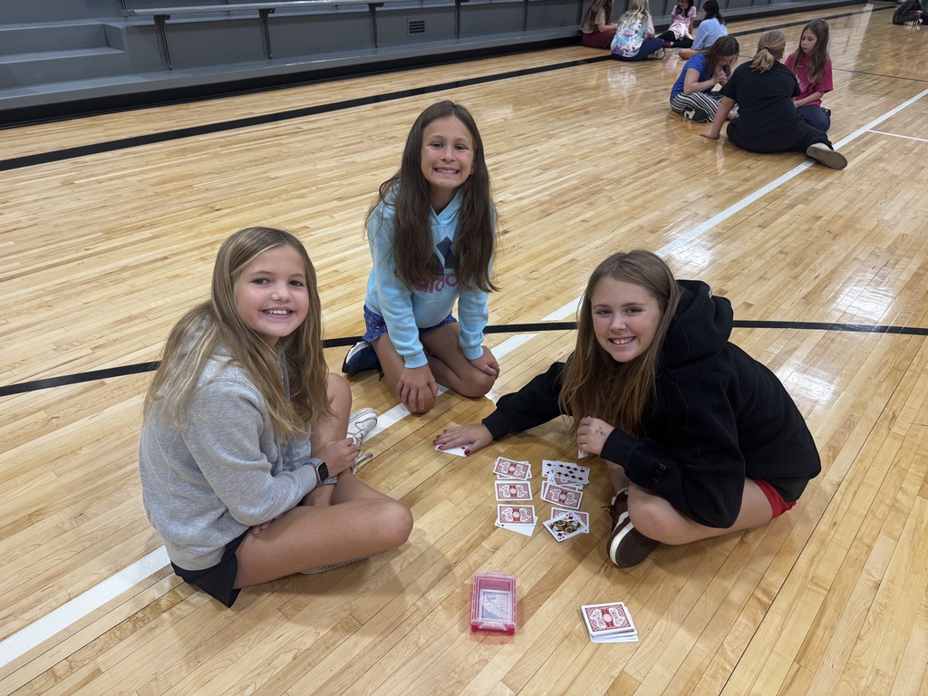 Three girls sitting on the gymnasium floor gathered around a deck of playing cards.