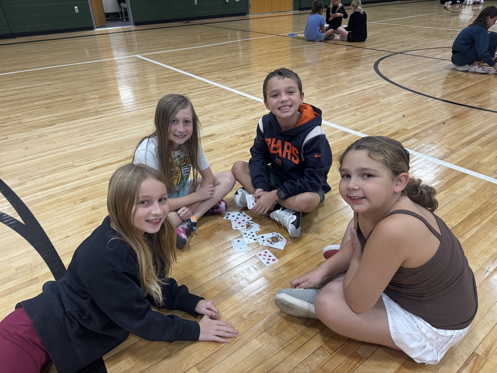 Four kids sitting cross-legged in a small circle on the basketball court playing cards.