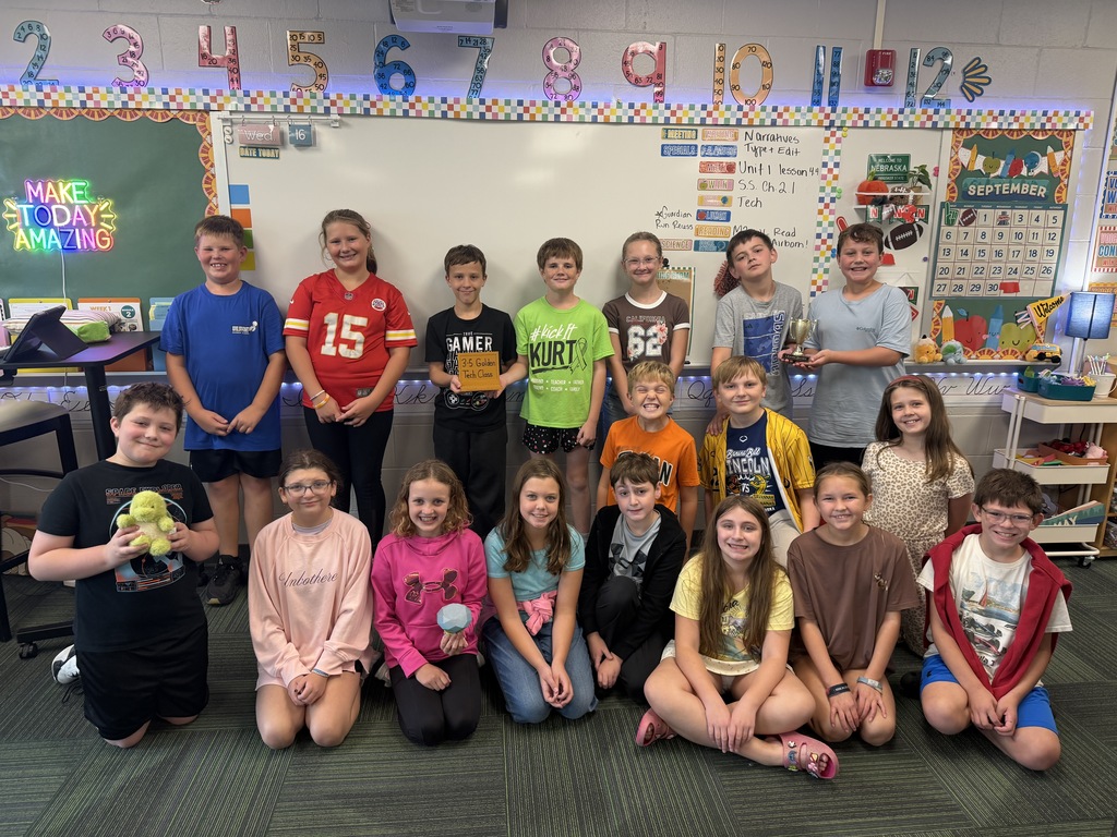 A classroom full of elementary students smiling as a group while displaying their trophies and awards in front of the whiteboard.
