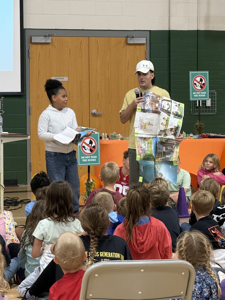 A guest speaker holding up artwork alongside a student while speaking into a microphone.