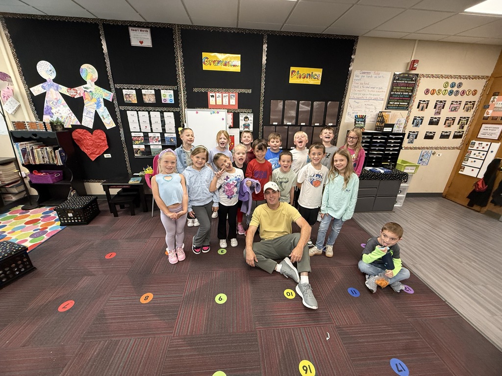 A guest presenter crouching in front of a group of smiling elementary students in a decorated classroom.