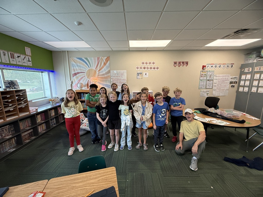 A guest speaker kneeling beside a group of elementary students posing together in their classroom.