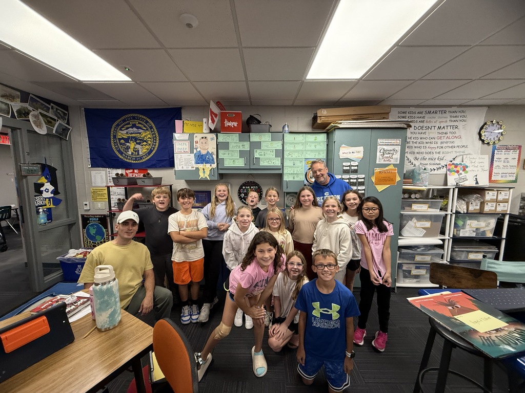 A classroom of students and teachers gathering around a desk for a group picture.