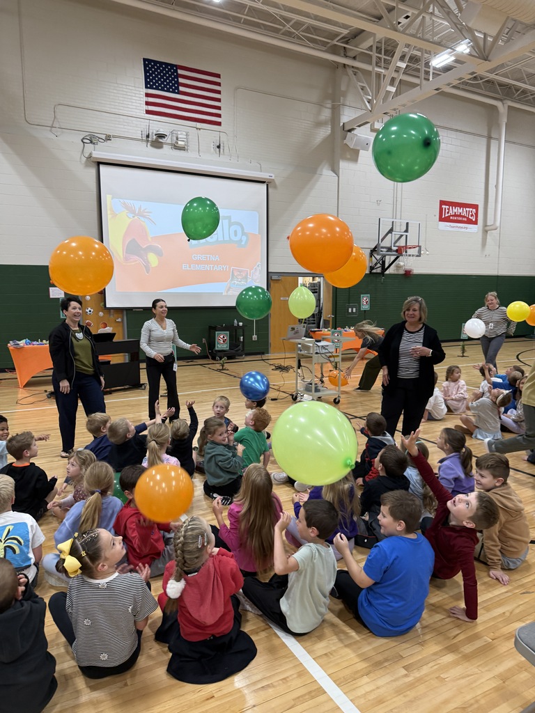Elementary students sitting on the gymnasium floor watching colorful balloons float down during an assembly.