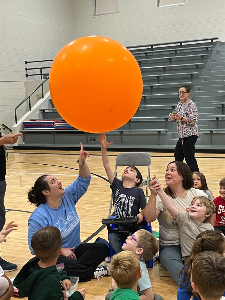 Teachers and a student reaching up to touch a large orange balloon floating in the gym.