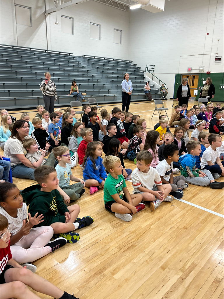 A wide view of students seated on the gymnasium floor during a presentation while staff members stand nearby.