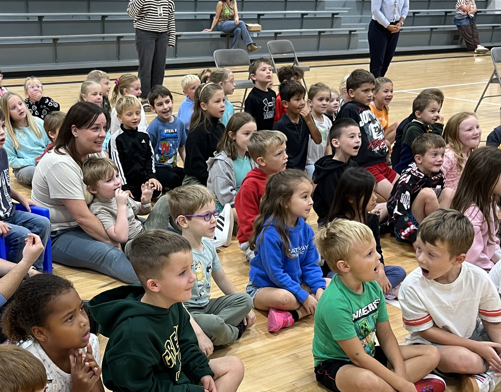 Elementary students sitting cross-legged on the gymnasium floor listening attentively during an assembly.