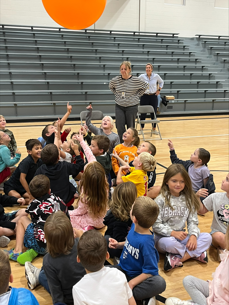 Students looking up and reaching toward a large orange balloon floating overhead in the gym.