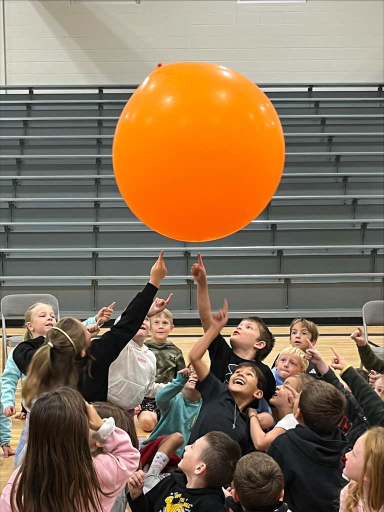 A group of kids reaching up with their fingers toward a giant orange balloon floating above them.