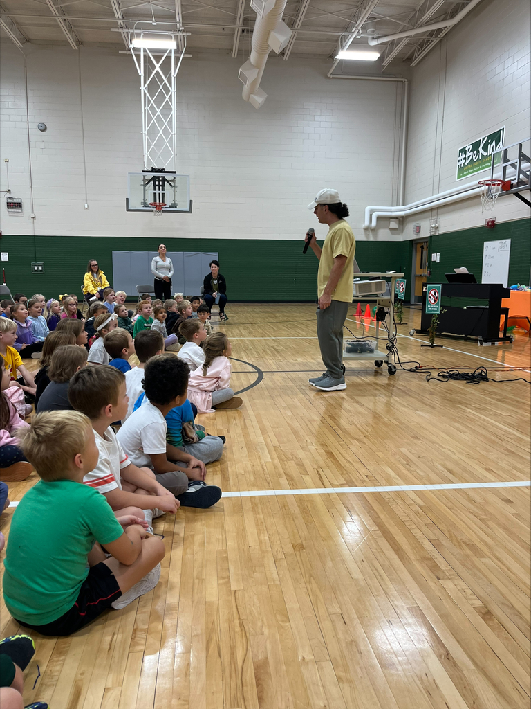 A presenter speaking into a microphone while standing in front of students seated on the basketball court.