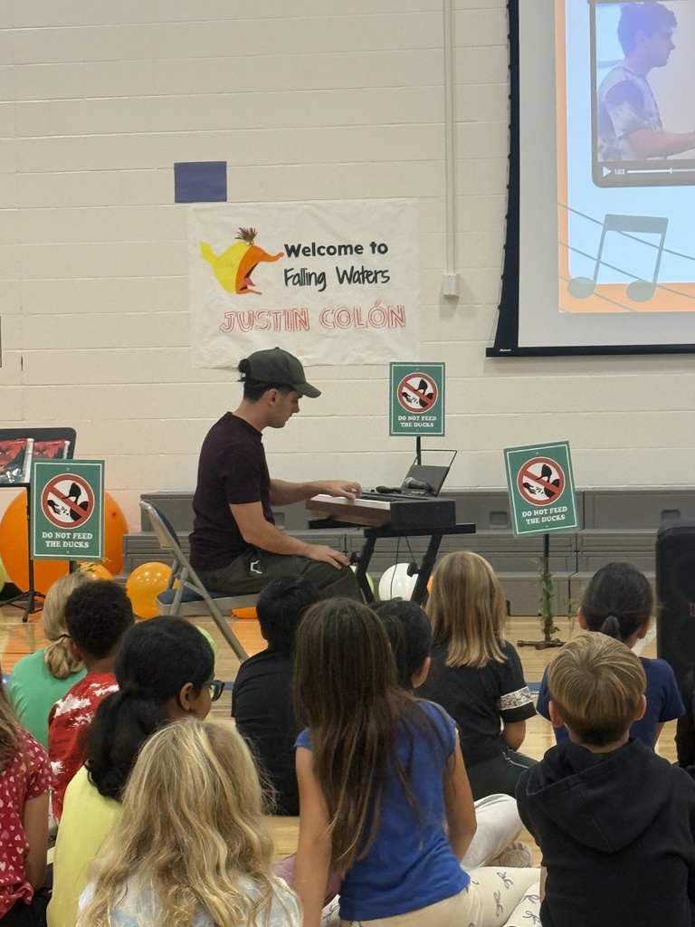 Justin is sitting at a piano while students sit in front of him watching.