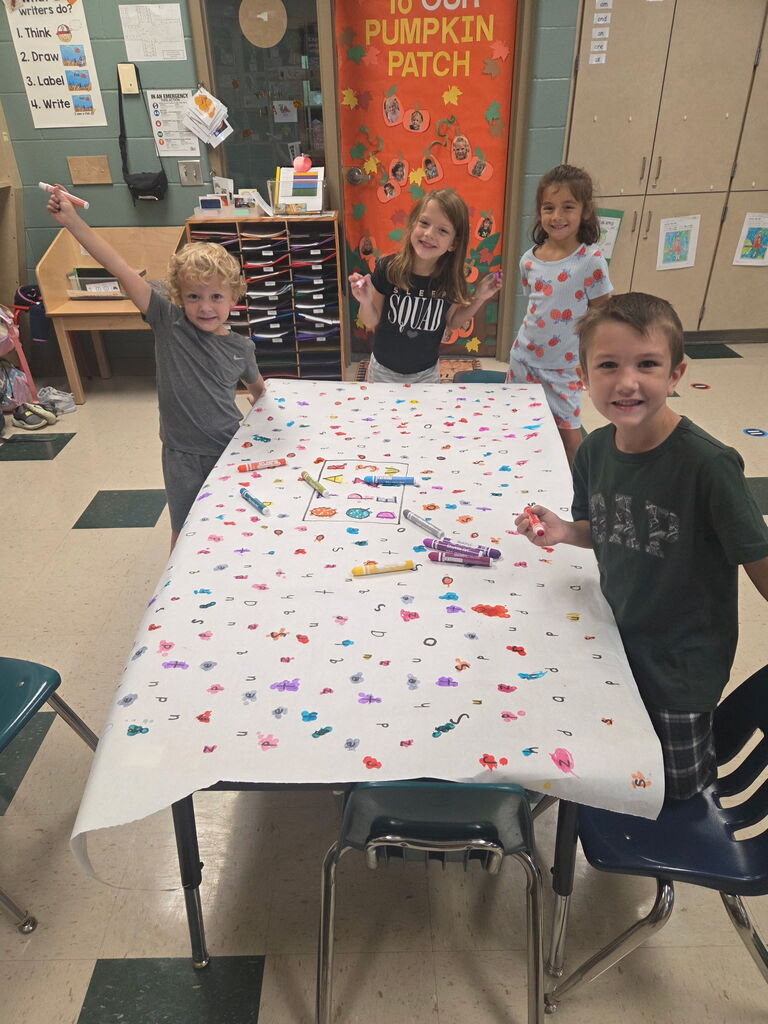 4 students are standing around a table with letters written on the tablecloth.