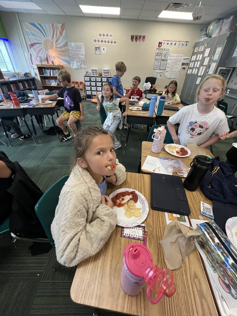 A student in a fuzzy jacket taking a bite of a snack while seated at her desk in the classroom.