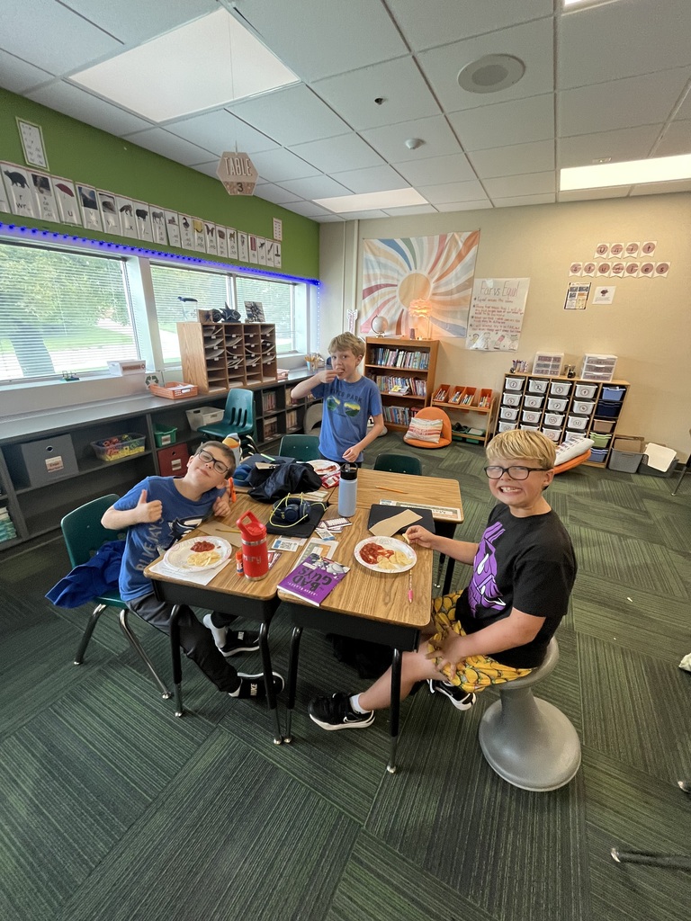 A group of young students sitting at connected desks giving thumbs-up gestures and smiling during snack time.