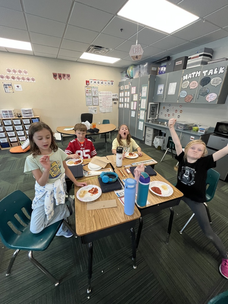 Elementary students at their desks eating paper-plate snacks, with one student playfully raising her arms.