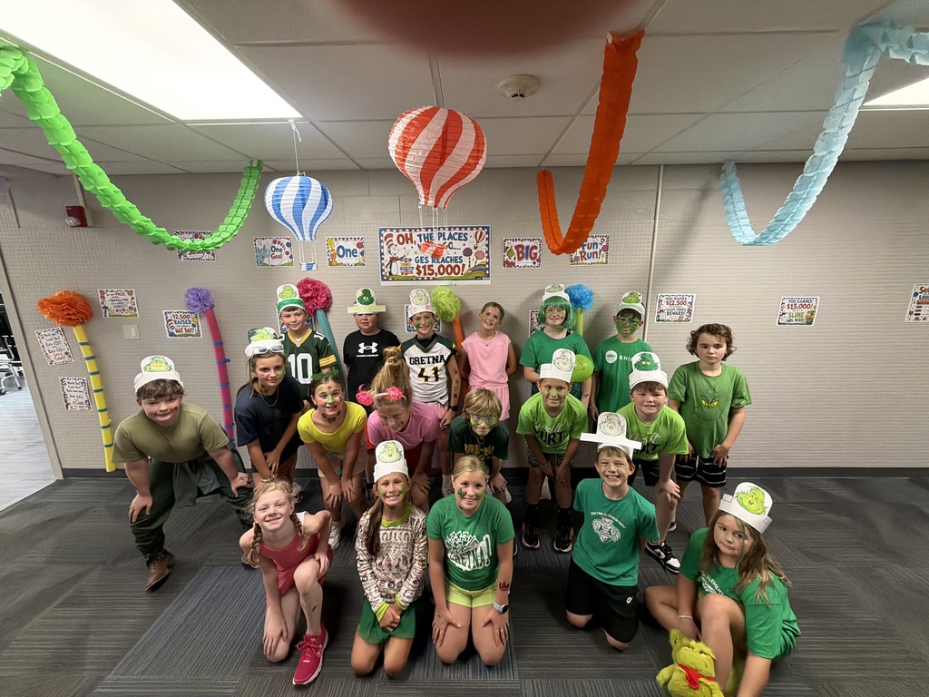 A group photo of students and teachers wearing green outfits and party hats under Dr. Seuss-themed classroom decorations.