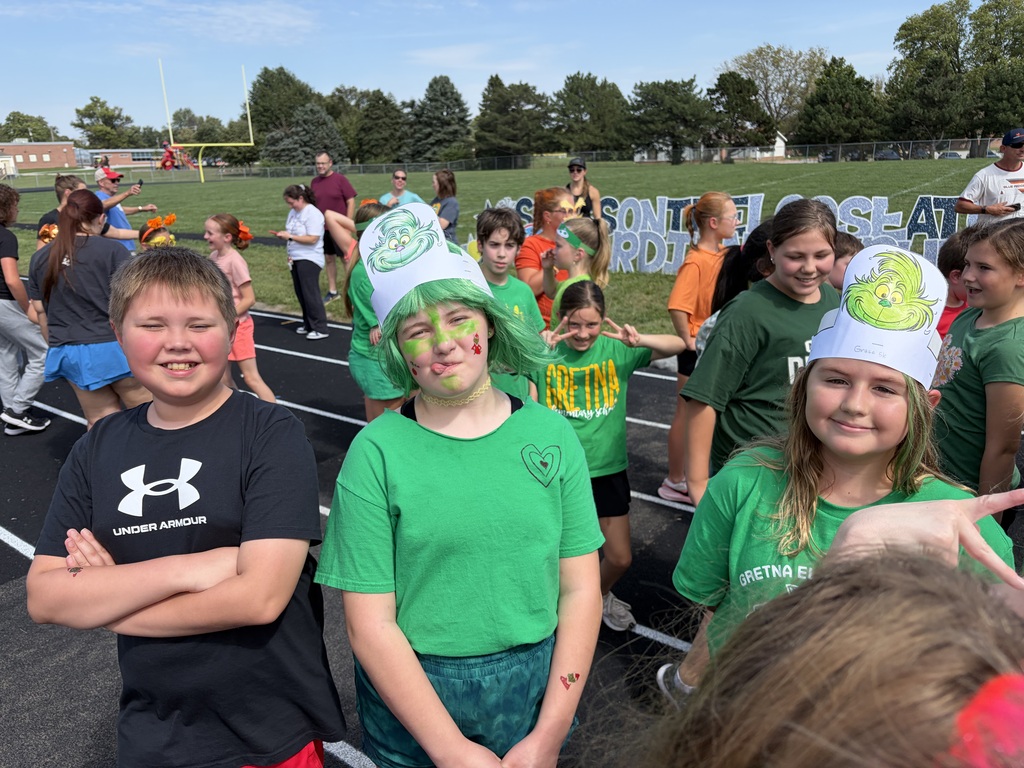 Students wearing green shirts and festive Grinch paper hats standing together outside near the track.