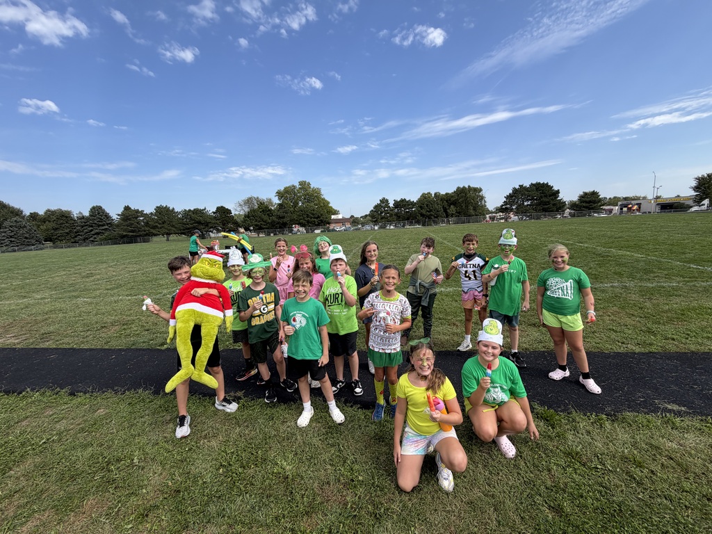 A large group of students in green gear gathering on the grass field with popsicles and a plush Grinch mascot.