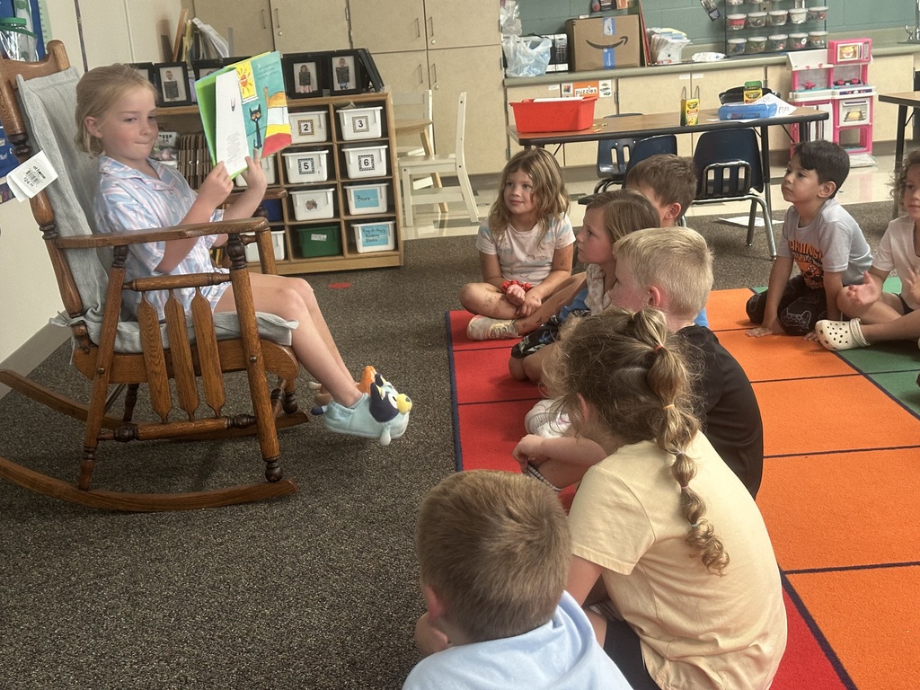 A student is in a rocking chair reading to students sitting on a rug.