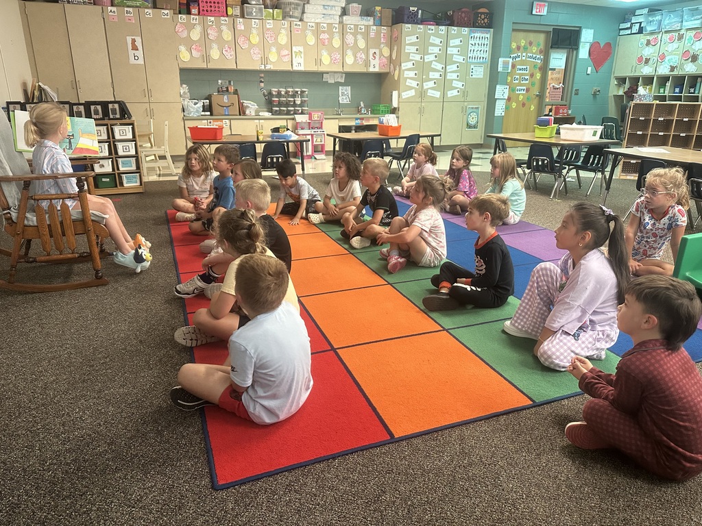 A student is in a rocking chair reading to students sitting on a rug.