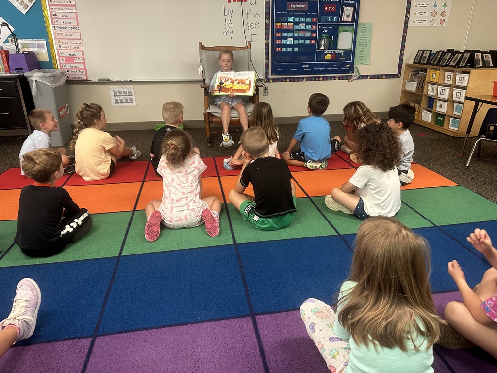 A student is in a rocking chair reading to students sitting on a rug.