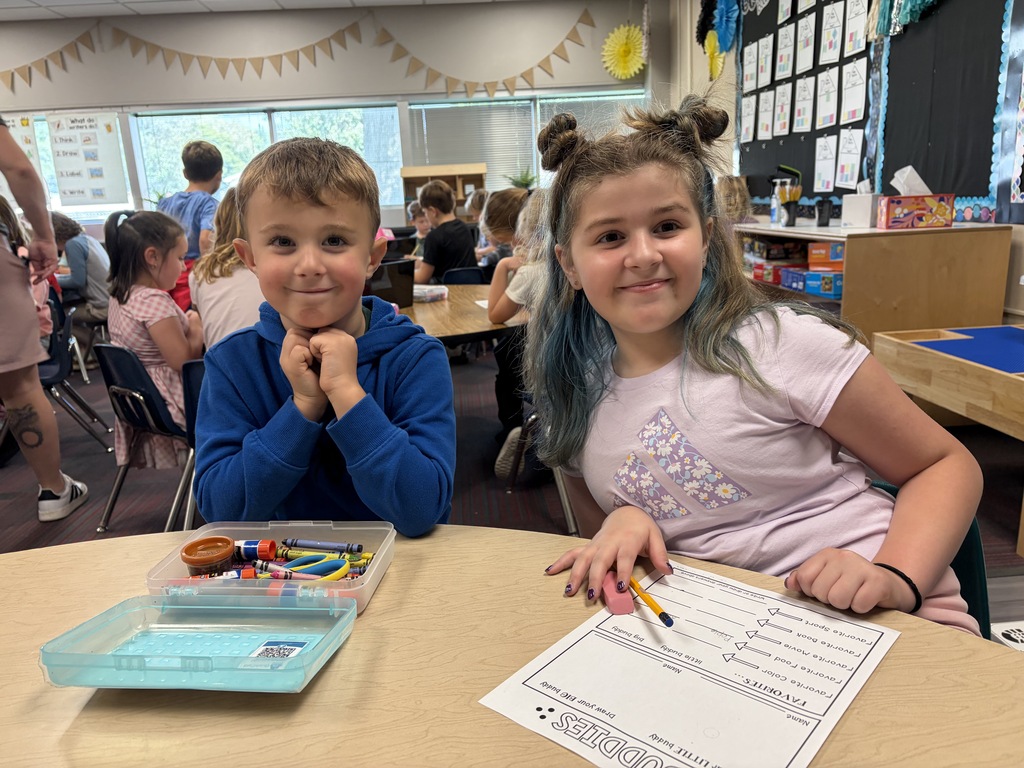 A boy in a blue hoodie and a girl with space buns sitting at a table with school supplies and a worksheet.