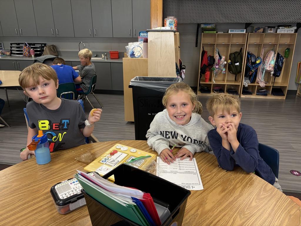 Three elementary students sitting around a wooden table in a classroom with "BUDDIES" worksheets and supply bins.