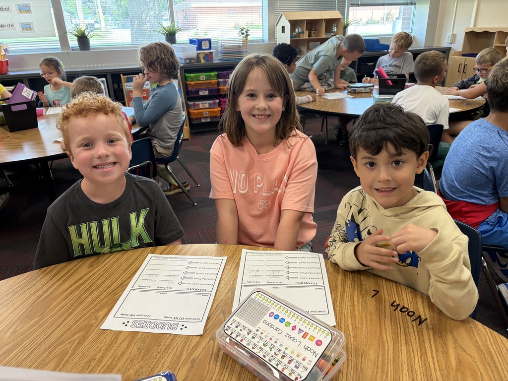 Three young students sitting side by side at a table with pencil boxes and "BUDDIES" worksheets in a classroom setting.